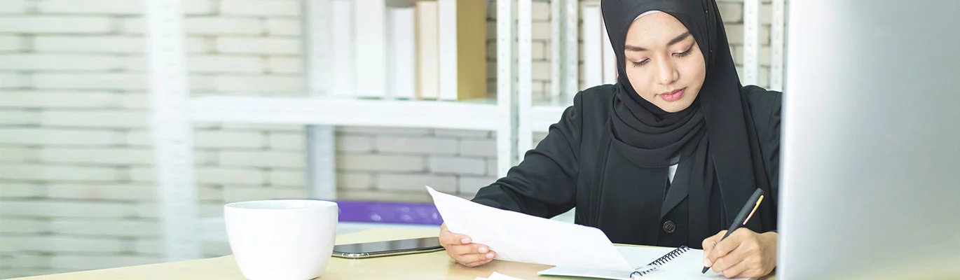 A student wearing purdah sitting in a private space holding a paper in one hand and copying on a book 