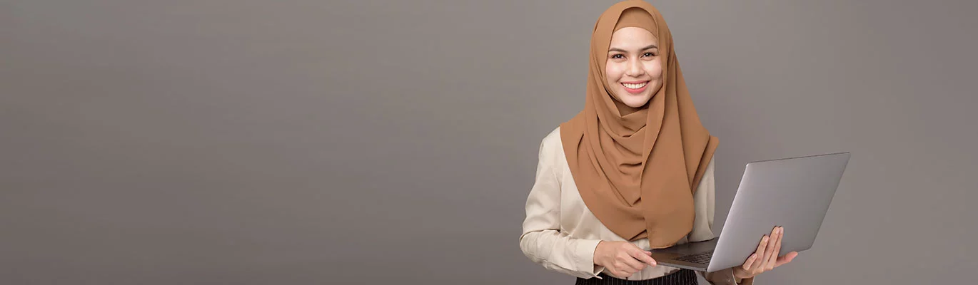 An Azure student wearing hijab standing in a ash color background with a laptop in hand & smiling at the camera