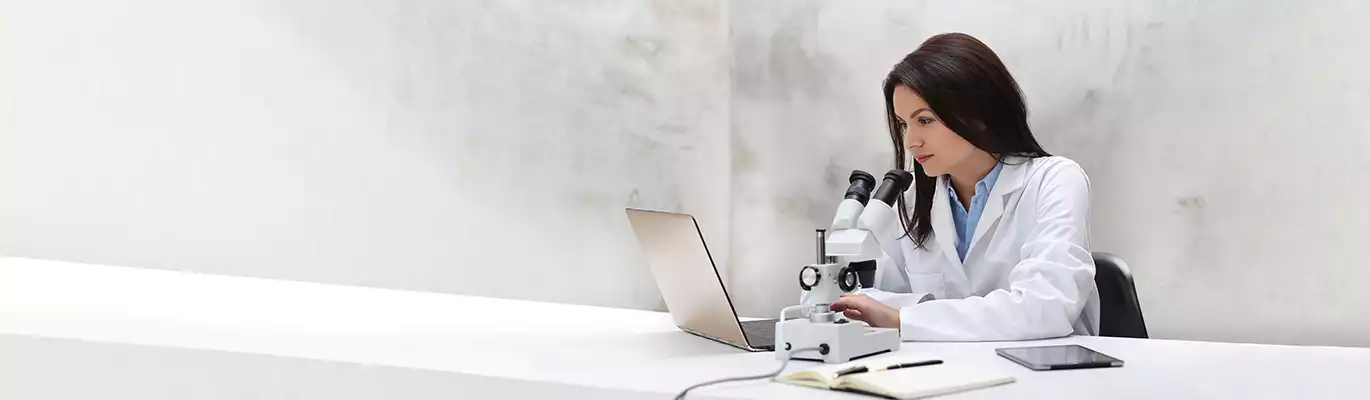 A Biologist in work unifrom sitting infront of a laptop and a microscope