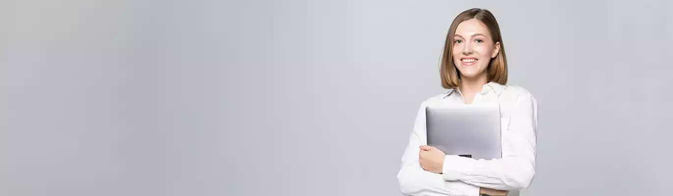 A professional woman in white shirt standing holding a folded laptop with a smiling face in a ash background