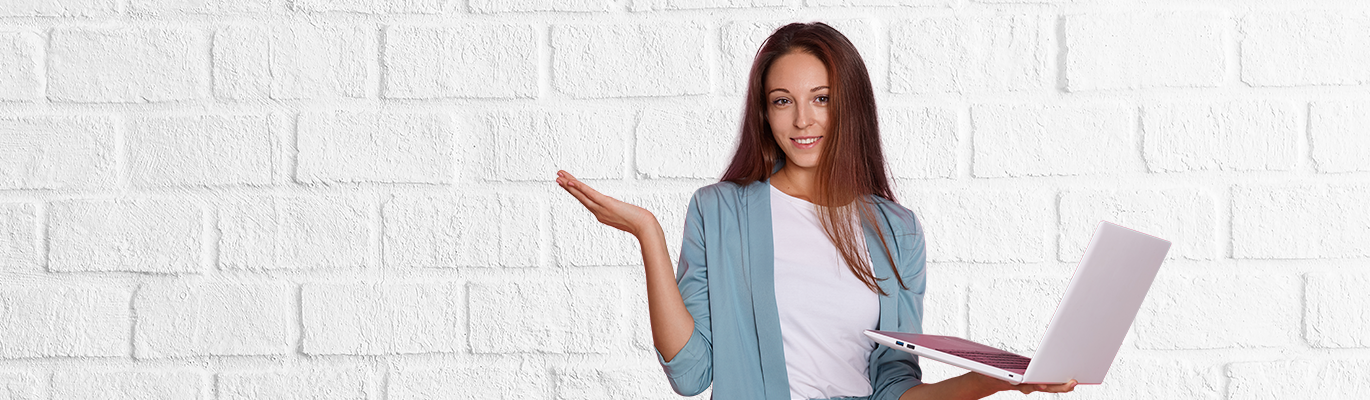 A SAP student standing in a brick wall background with a laptop in hand, smiling at the camera with an upwardopen hand gesture 