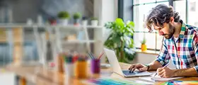 Man working on graphic design using laptop and color samples in a creative workspace.