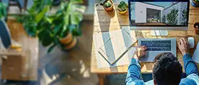 Man working on a laptop with architectural design on screen at a wooden desk with plants.