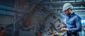Engineer in hard hat working on laptop in an automated industrial setting.
