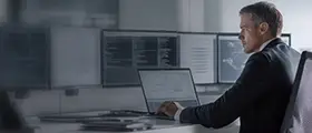 Professional man working on a laptop in a modern control room with multiple monitors displaying data and code.