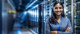A smiling woman stands with arms crossed in a server room, with rows of server racks behind her.