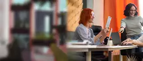Two women collaborating at a desk, discussing and pointing at a laptop screen.