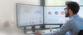 Man analyzing data and charts on dual monitors in an office environment.
