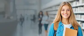 Smiling young woman holding a folder stands in a library or academic setting, with bookshelves and other students in the background.
