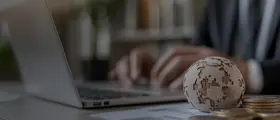 Person typing on a laptop with a small globe and stacked coins in the foreground, symbolizing global finance.