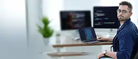 Developer working on laptop with code on multiple screens in modern office.