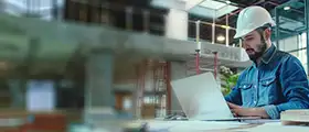 Engineer wearing a hard hat working on a laptop at a construction site.