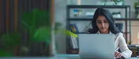 A woman with long dark hair is focused on a laptop screen while working at a desk.