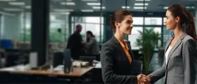 Two professional women shaking hands confidently in a modern office setting, symbolizing successful negotiation or business agreement.