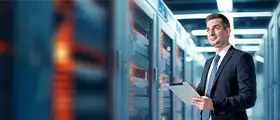 A smiling man in a suit and tie holds a tablet, standing in a server room with rows of glowing server racks.