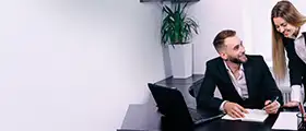 Two professionals discussing work at an office desk with a laptop.