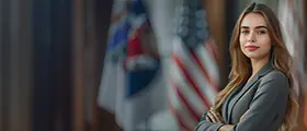 A confident woman in business attire stands with arms crossed in front of blurred U.S. and other flags, suggesting a professional or governmental setting.