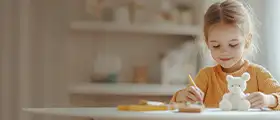 A young girl painting a white bear-shaped ceramic figure at a table, smiling and focused.