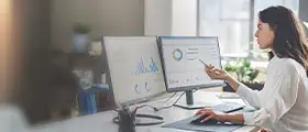 Professional woman analyzing data dashboards on dual monitors in a modern office.