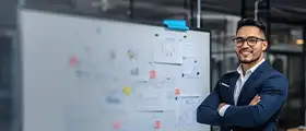 A smiling man with glasses and a dark suit stands with his arms crossed in front of a whiteboard covered with notes and diagrams.