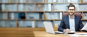 A businessman in a suit working at a desk with a laptop and documents, sitting in front of a bookshelf.