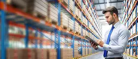 Man using a tablet in a warehouse aisle with shelves stacked with boxes.