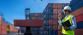 A worker in a safety vest and helmet communicates via walkie-talkie at a busy shipping yard with stacked cargo containers.