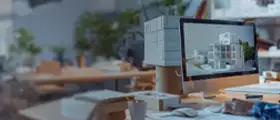 A modern office workspace with a computer displaying a 3D architectural model, surrounded by papers, books, and design materials.