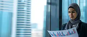 Businesswoman in hijab analyzing a printed financial chart by a window with a cityscape in the background.