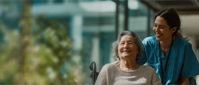 A professional caregiver assisting an elderly woman in a wheelchair, smiling together in a bright healthcare setting, representing compassionate elderly care and caregiver training.