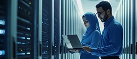 Two IT professionals analyzing data on a laptop inside a modern server room with network racks and glowing equipment.