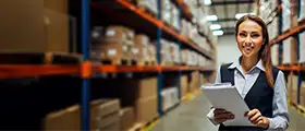 Certified Warehouse Management Professional standing with a clipboard in a large warehouse filled with shelves of boxes, smiling confidently.