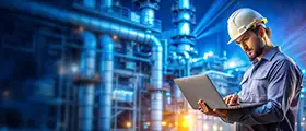 A professional engineer wearing a safety helmet using a laptop in an industrial facility, representing competency assurance and management systems.