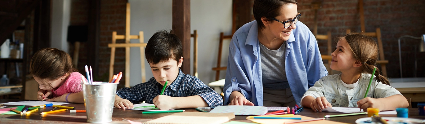Two children drawing when the teacher smilingly interacts with another child who is also drawing
