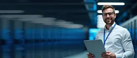 IT professional holding a laptop and smiling confidently inside a modern data center, with rows of illuminated server racks in the background.