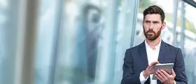 Business professional in a suit using a digital tablet while standing in a modern glass-walled office corridor, symbolizing performance management and strategic decision-making.