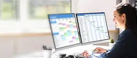 A professional project scheduler working on dual monitors displaying project timelines and calendars for scheduling management.