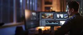 A professional video editor working on multiple monitors displaying video editing software in a dimly lit creative studio.