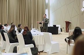 Professional corporate trainer delivering a presentation to a seated audience in a conference room, with attendees listening attentively during a business seminar or workshop.